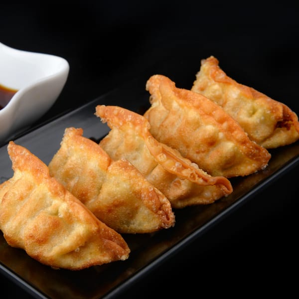 Five golden-brown fried dumplings, or gyoza, arranged on a black rectangular plate with a white sauce dish.