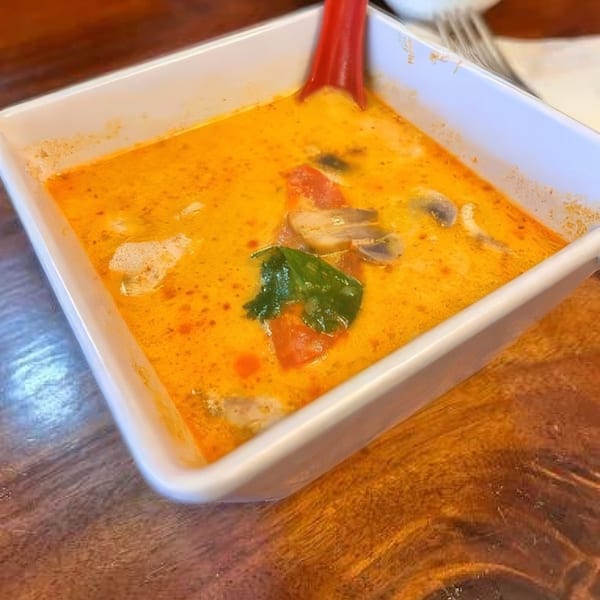 A white square bowl of orange Tom Yum soup with mushrooms, tomatoes, and a green leaf.