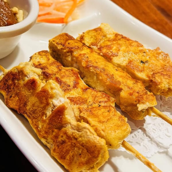 Three golden-brown chicken satay skewers on a white plate with peanut sauce and pickled vegetables.